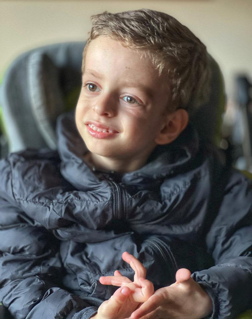 Théo, de oito anos, sorrindo para foto. Veste casaco preto e está sentado em sua cadeira