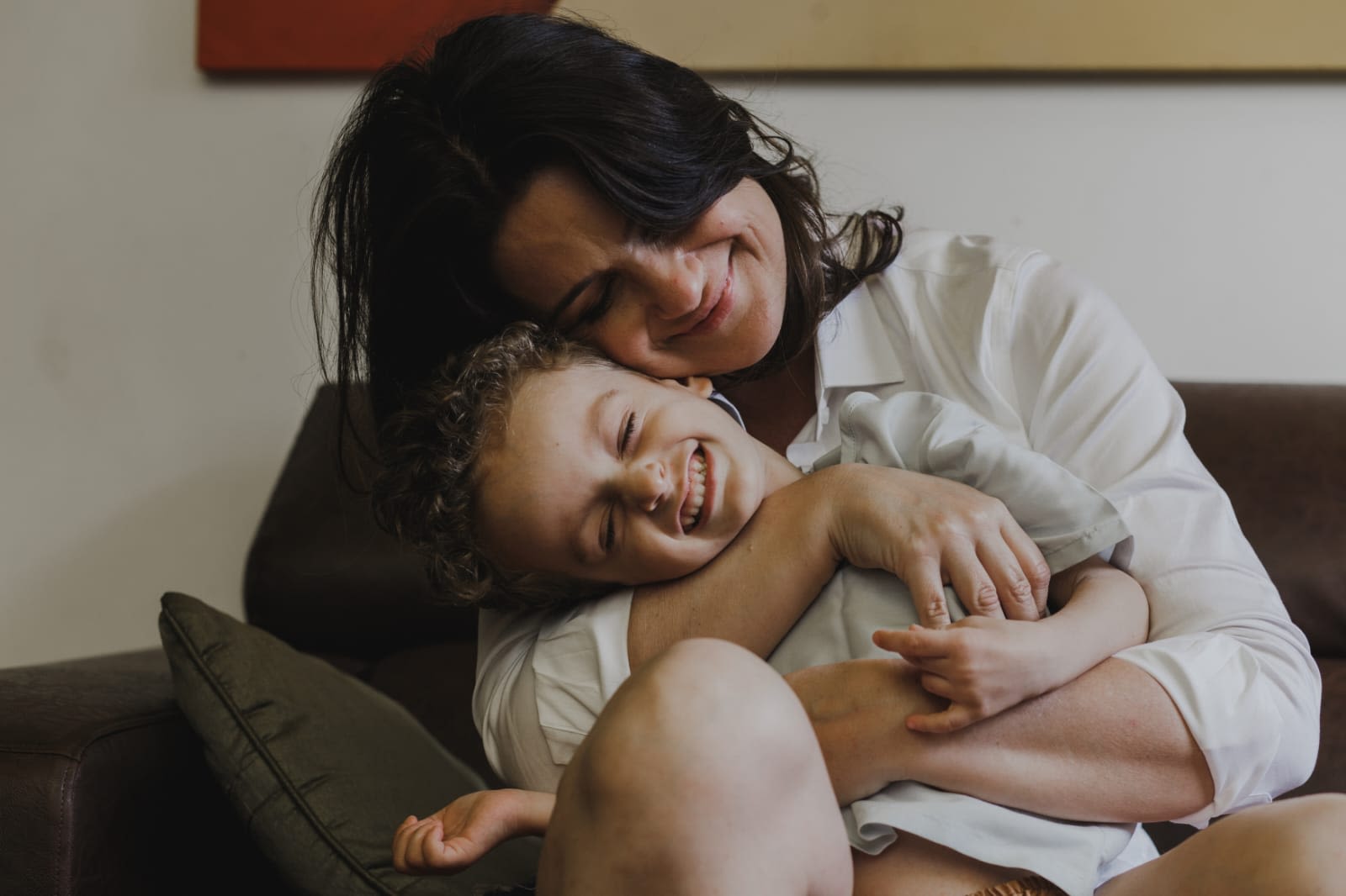 Mãe e filho abraçados, usando roupas brancas e sentados num sofá verde musgo. Ambos sorriem. A mãe é branca, de cabelos pretos lisos. O filho também é branco, e tem cabelos loiros chacheados
