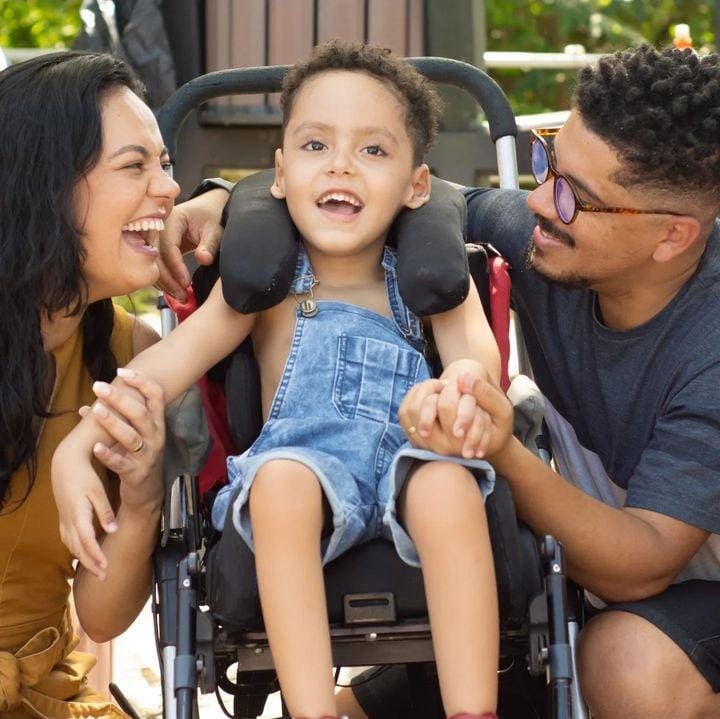 Natalia, Matias e Marlon posando para foto. Os pais estão sorrindo para Matias, centralizado na foto. O menino, que veste macacão jeans e está sentado em sua cadeira, também sorri, olhando para a câmera.