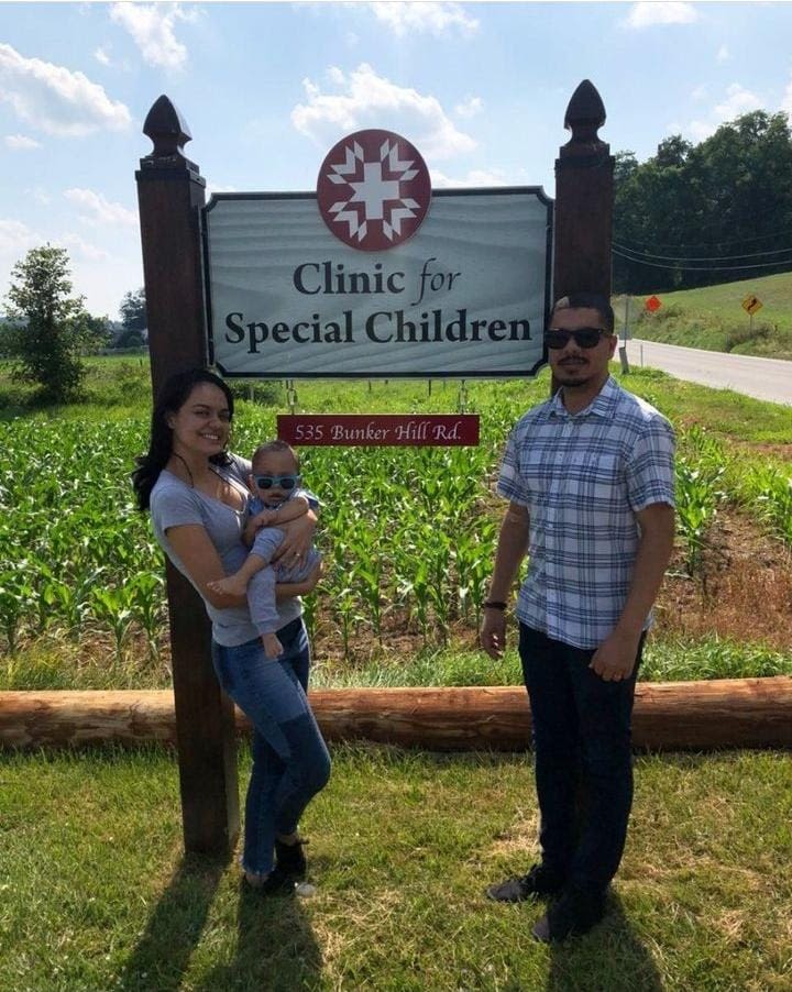 Nathalia e Marlon posando em frente a placa da Clinic for Special Children, com um campo como paisagem atrás. Ambos vestem jeans e Marlon usa óculos de sol e camisa azul quadriculada. Nathalia segura Matias no colo, e a criança também está de óculos. A mãe usa blusa cinza
